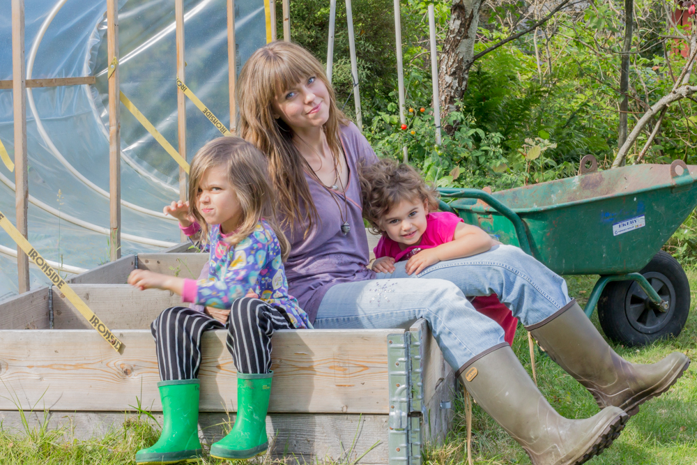 Mamma med döttrar sittandes i en pallkrage bland odlingar i trädgård.